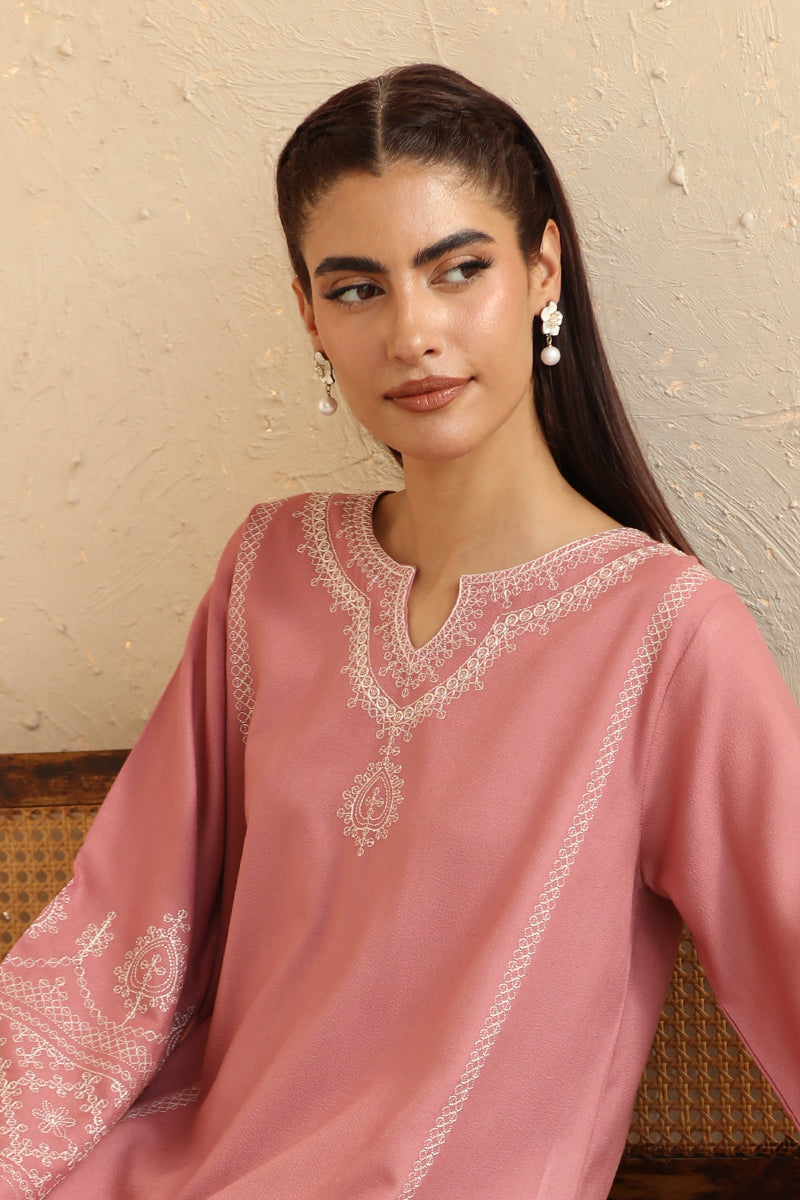 Woman wearing a pink embroidered traditional outfit against a beige wall.