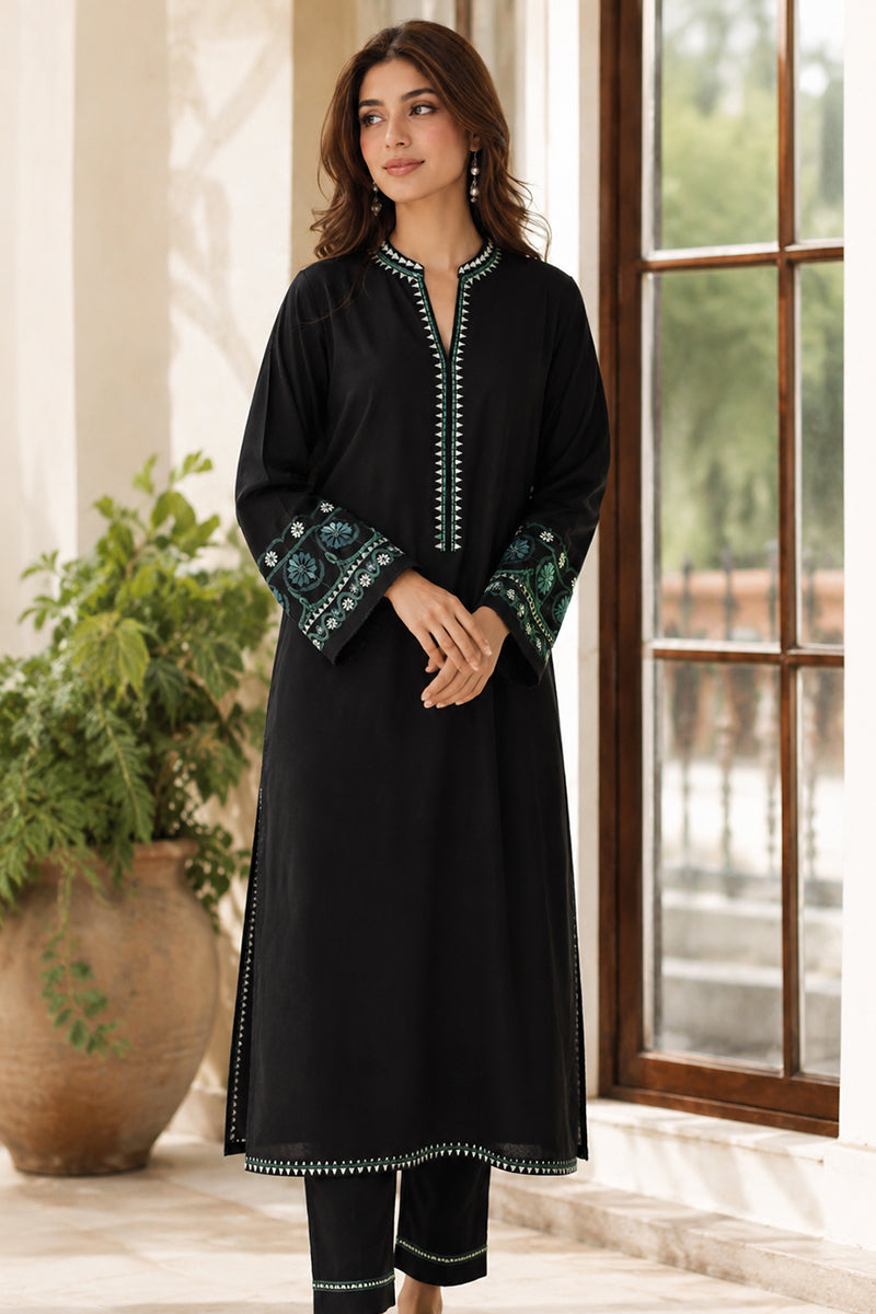 Woman wearing a black traditional outfit with green embroidery in a room with a window and plant.