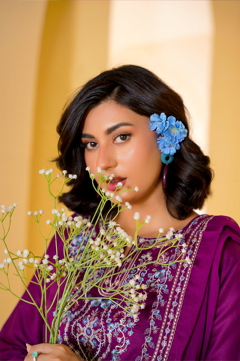 Woman in a purple traditional outfit holding flowers with a blue flower in her hair against a beige background