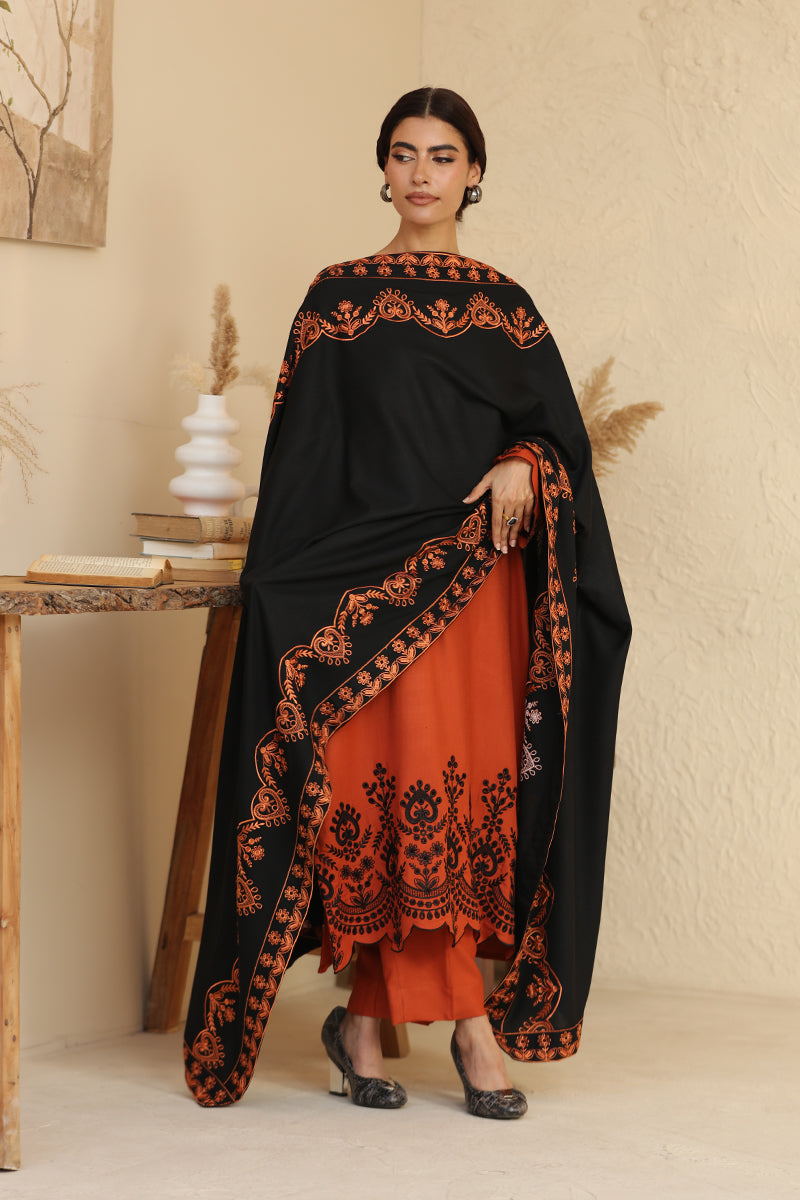 Woman wearing a black and orange traditional outfit with intricate patterns in a minimalistic setting.
