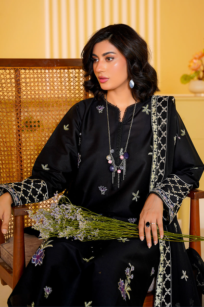 Woman in a black embroidered outfit holding flowers, sitting on a wooden chair.
