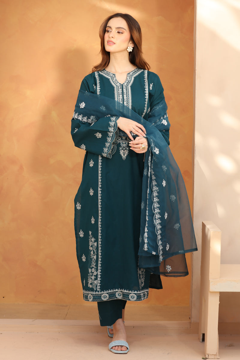 Woman wearing a teal embroidered outfit against a beige wall.