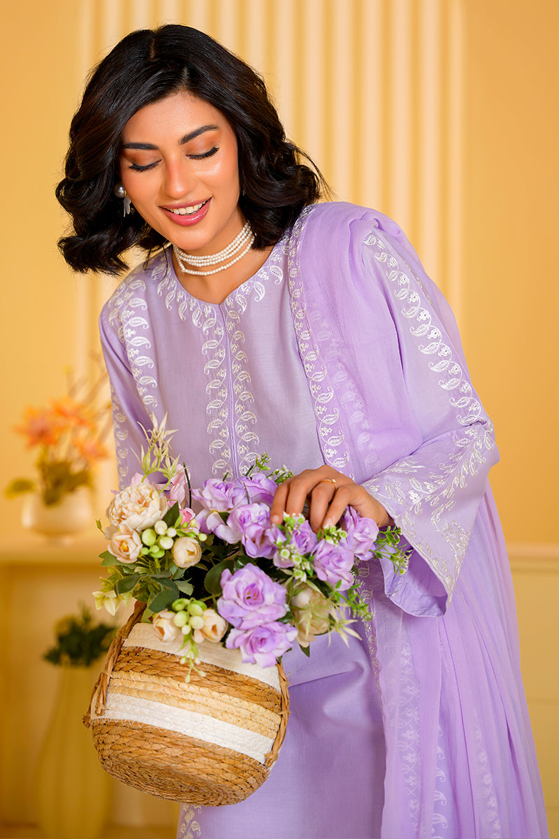 Woman in a lavender traditional outfit holding flowers and a woven basket against a yellow wall.