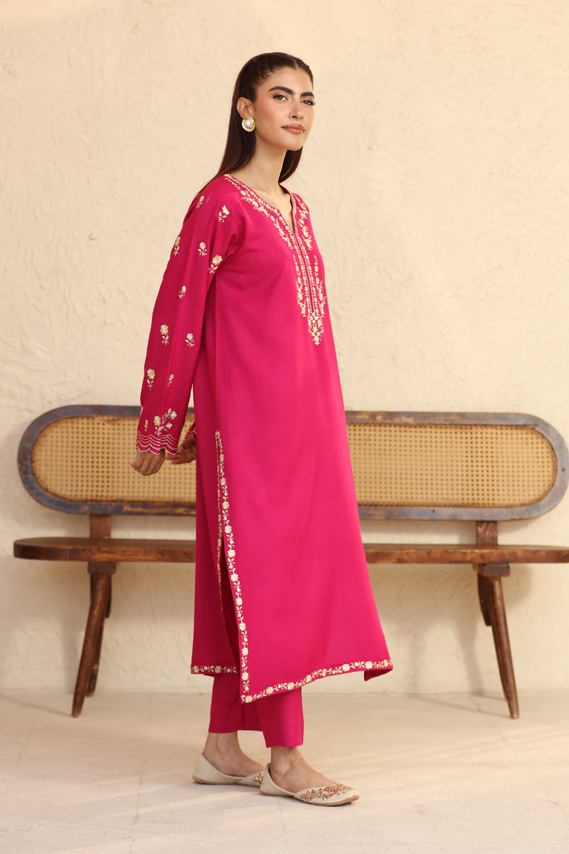 Woman wearing a pink traditional outfit with white embroidery, standing against a beige wall.
