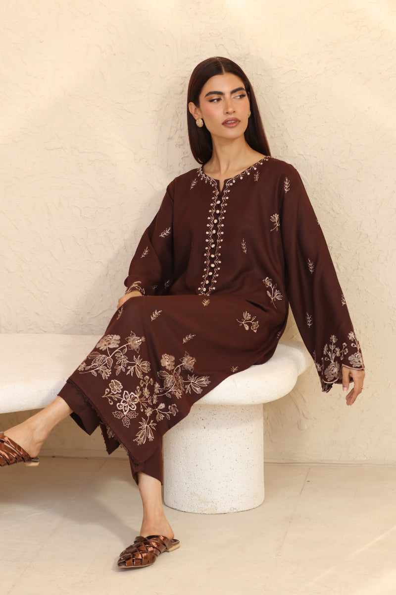 Woman wearing a brown embroidered outfit sitting on a white chair against a beige wall.