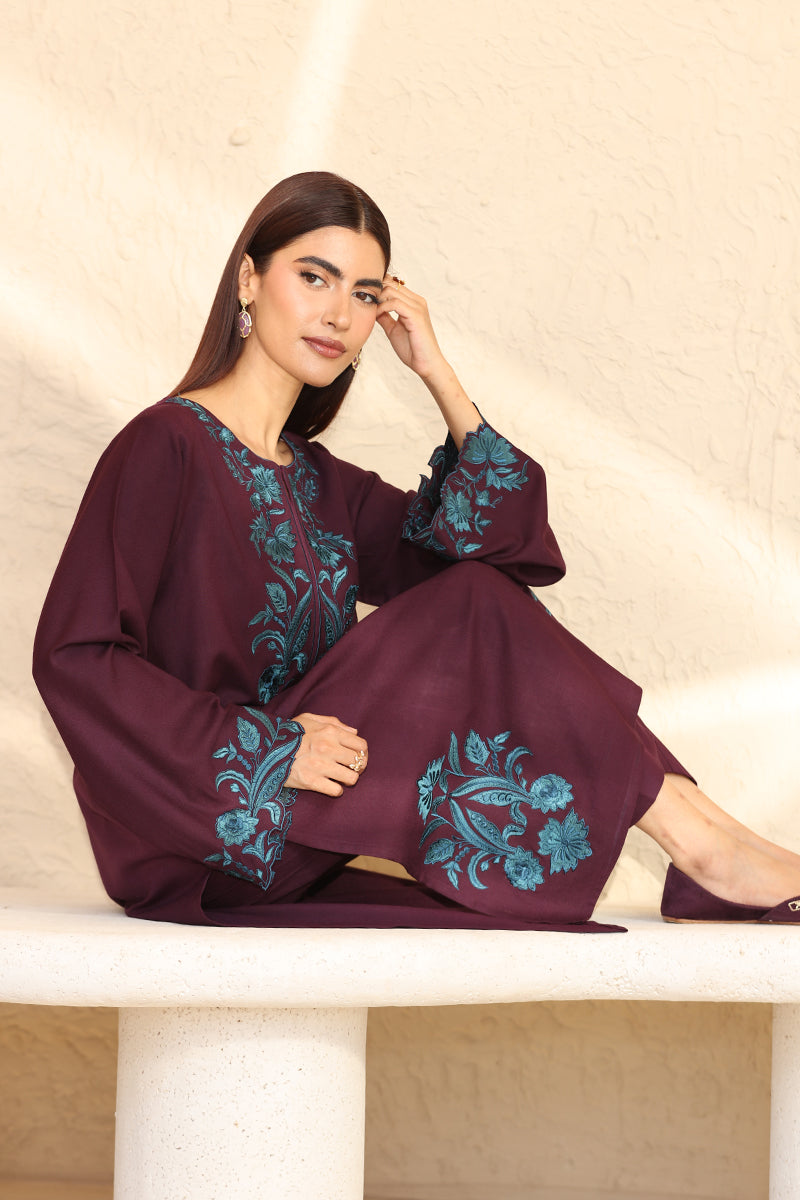 Woman wearing a burgundy dress with blue floral embroidery sitting on a white surface.