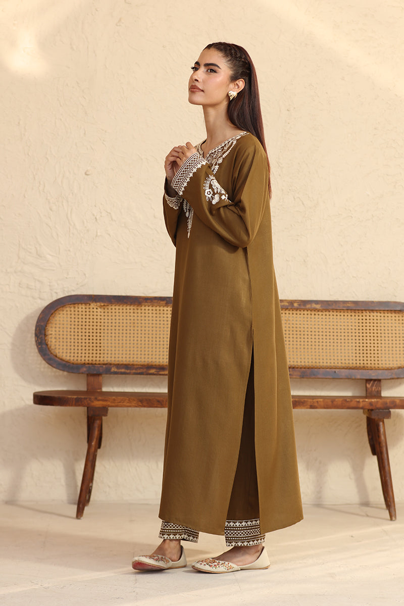 Woman wearing a long olive green dress with white patterns, standing against a beige wall with a wooden bench.