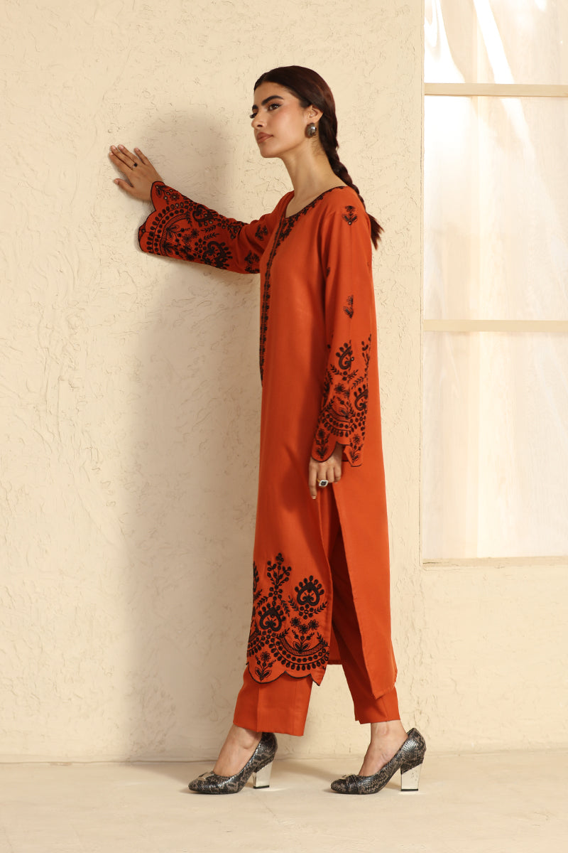 Woman wearing an orange outfit with black embroidery against a beige wall.