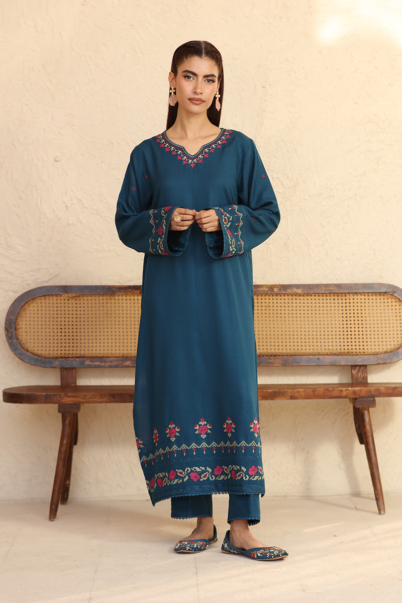 Woman wearing a blue embroidered traditional outfit standing in front of a wooden bench.