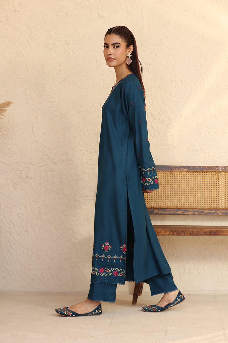 Woman wearing a teal embroidered outfit standing against a beige wall.