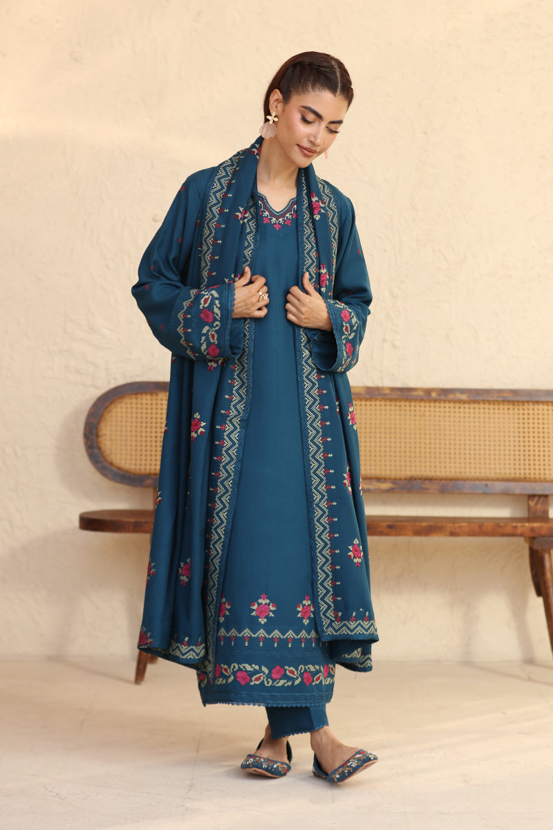 Woman wearing a blue embroidered outfit standing in a minimalistic room.