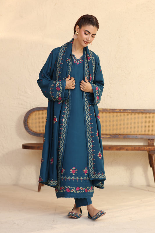 Woman wearing a blue embroidered outfit standing in a minimalistic room.